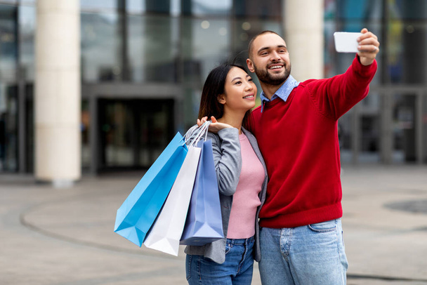 Retrato de una joven pareja multirracial haciendo selfie para teléfonos inteligentes, sosteniendo bolsas de regalo brillantes después de comprar en el centro comercial de la ciudad - Foto, Imagen