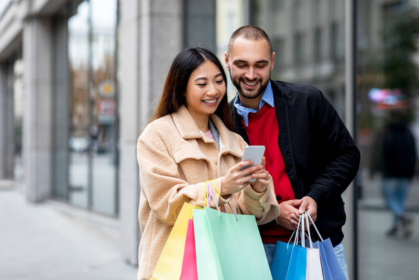 Positives junges, vielseitiges Paar mit hellen Papiertüten und Handy, das in der Nähe von Einkaufszentrum Waren online bestellt - Foto, Bild