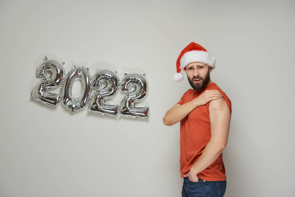 Een verraste kale man met een baard in een kerstmuts toont gips op zijn schouder na vaccinatie tegen het Coronavirus (Covid-19) in de buurt van verijdelde zilveren ballonnen in de vorm van 2022. - Foto, afbeelding