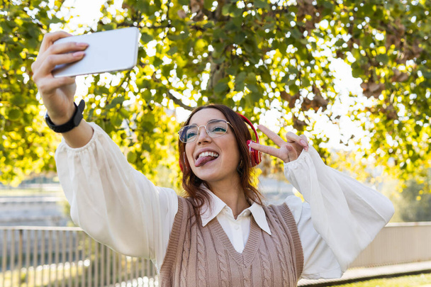 Attraktive junge rothaarige Geschäftsfrau mit Brille, die die Zunge herausstreckt und Spaß beim Selfie mit dem Smartphone und beim Musikhören im Freien hat - Foto, Bild