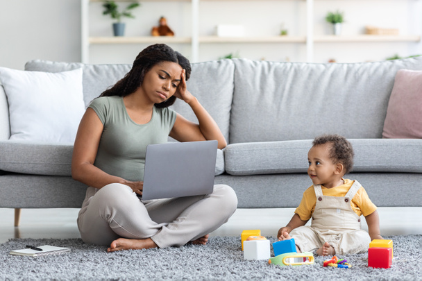 Измученная черная мать пытается работать на ноутбуке с маленьким ребенком вокруг - Фото, изображение