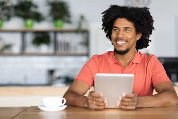 Glücklicher schwarzer junger Kerl entspannt sich im Cafe mit digitalem Tablet und Kaffee - Foto, Bild