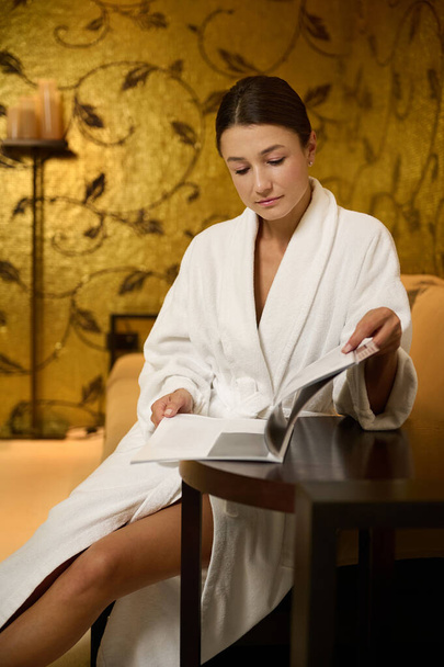 Confident attractive relaxed young woman resting in a lounge zone of a wellness spa clinic, wearing bathrobe, reading magazine and getting ready for receiving professional body and skin care treatment - Valokuva, kuva