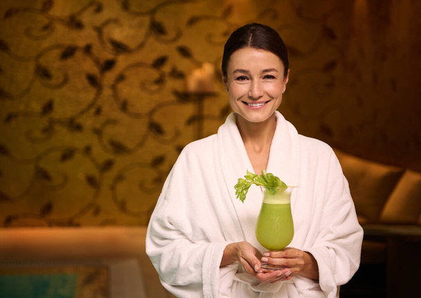 Charming middle aged European woman in white terry bathrobe, holding a glass with freshly squeezed vitamin juice in her hands, smiles toothy smile looking at camera while relaxing at wellness spa - Foto, Imagem