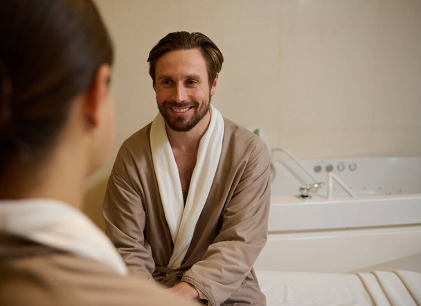 Handsome middle aged European man in bathrobe smiling looking at his wife, out of focus on the foreground, resting at wellness spa resort on honeymoon. Cute couple getting beauty treatment together - Photo, Image