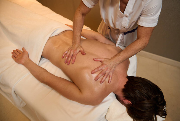 Visão aérea do terapeuta de massagem profissional irreconhecível massageando os músculos traseiros de um jovem, relaxante, deitado na mesa de massagem no centro de spa de bem-estar de luxo. Conceito de terapia alternativa - Foto, Imagem