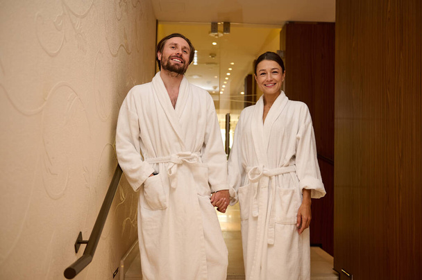 Beautiful loving newlywed couple, wearing white bathrobes, enjoying their honeymoon together in luxury spa resort, holding hands as they walk down the stairs of the wellness spa complex - Photo, Image