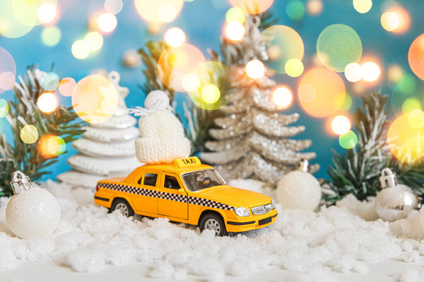 Kerstspandoek Achtergrond. Gele speelgoedauto Taxi Cab model en winter decoraties ornamenten op blauwe achtergrond met sneeuw en onscherpe slinger verlichting. Stadsverkeer levering taxi service concept. - Foto, afbeelding