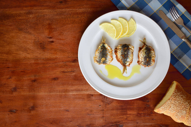 Background of a wooden table top with a plate of baked sardines on it, with bread, cutlery and a napkin on the edge, with space for text - Photo, Image