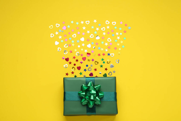Green gift box and shiny confetti on yellow background, top view - 写真・画像