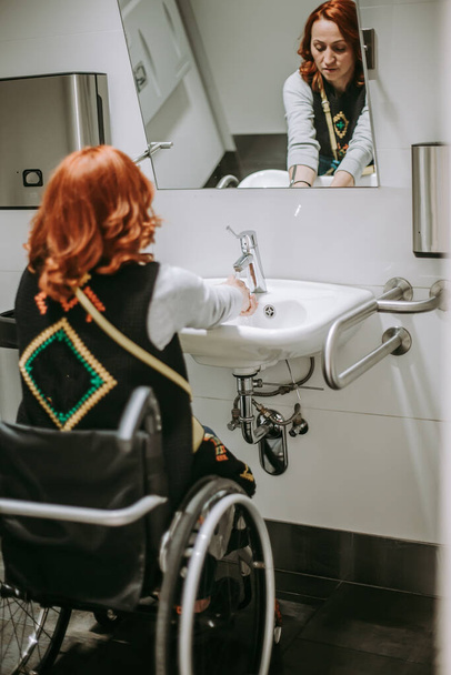 車椅子を必要とする女性は公衆トイレで手を洗う。車椅子の人が利用できる特別なニーズを持つ人々のためのトイレ。障害者用トイレ - 写真・画像