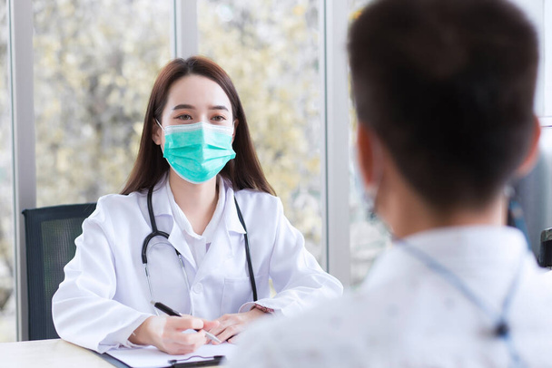 Jeune femme asiatique professionnelle médecin porte manteau médical et masque facial lors de l'examen et parle avec un patient homme dans la salle d'examen à l'hôpital. - Photo, image