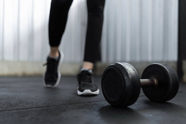 exercise concept The black rubber coated round dumbbell lying on the gym ground in front of the person who wears black trainers. - Foto, Imagem