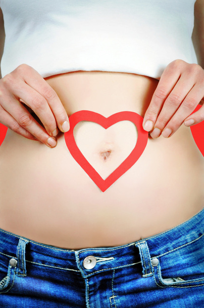 Gros plan portrait d'une jeune femme tenant une forme de coeur rouge sur le ventre autour du nombril. Fond rouge
. - Photo, image
