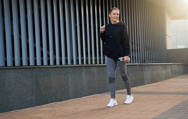 Full length shot of cheerful middle aged sportswoman smiling away while walking in the city, ready for workout outdoors - Fotó, kép
