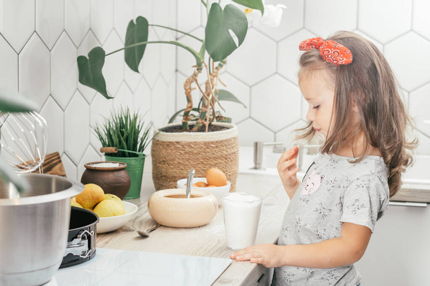 Little dark-haired girl 3 years old in red headband bakes apple pie in kitchen. Child puts sugar in measuring cup. Children help on household chores. Kid cooking food - Photo, Image