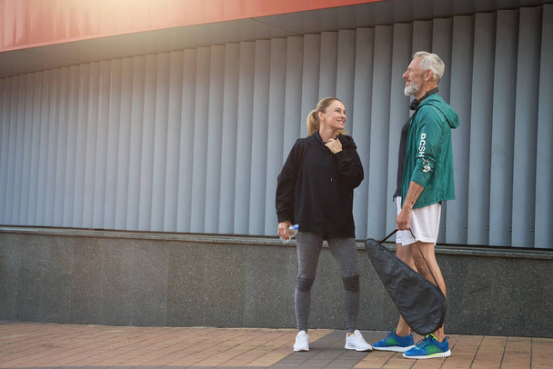 Cheerful middle aged sportspeople, man and woman in sportswear talking while standing together outdoors ready for workout - 写真・画像