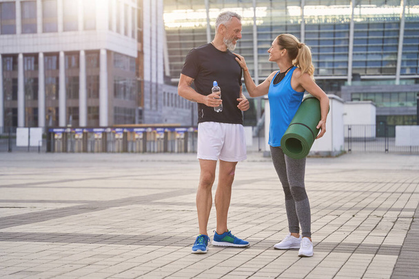 Ativo casal de meia-idade em sportswear fazendo esportes juntos na cidade, de pé ao ar livre com tapete de ioga pela manhã - Foto, Imagem