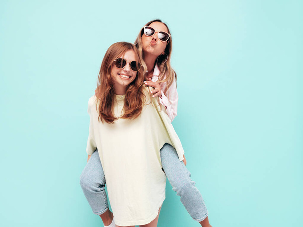 Two young beautiful smiling brunette hipster female in trendy summer clothes. Sexy carefree women posing near blue wall. Positive models having fun in sunglasses. Gives piggyback riding  - Fotó, kép