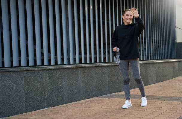 Pełna długość ujęcia sportowej kobiety w średnim wieku w odzieży sportowej uśmiechającej się i machającej podczas spaceru na świeżym powietrzu po treningu - Zdjęcie, obraz