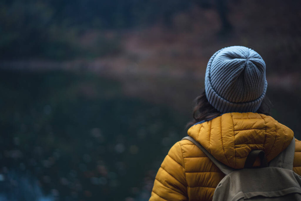 Młoda kobieta podróżuje w żółtej kurtce i kapeluszu stojąc plecami w mglistym lesie. - Zdjęcie, obraz