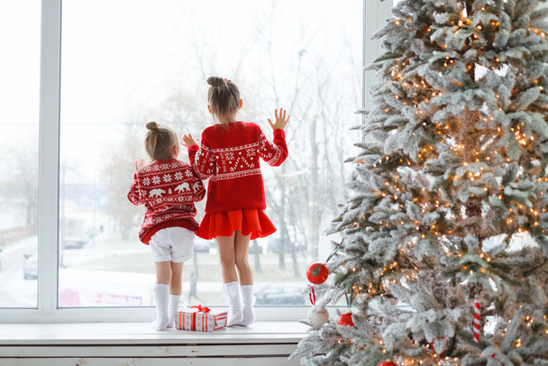 Children are standing on the windowsill with their hands on the glass and looking out the window impatiently waiting for Christmas and watching the falling snow outside. Contour soft light.red clothes - Photo, Image