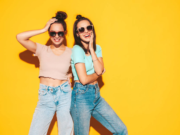 Two young beautiful smiling blond hipster female in trendy summer clothes. Sexy carefree women posing near yellow wall in studio. Positive models having fun. Cheerful and happy. In sunglasses - Foto, afbeelding