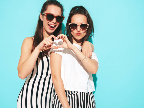 Two young beautiful smiling blond hipster female in trendy summer clothes. Sexy carefree women posing near blue  wall in studio. Positive models making heart sign. Cheerful and happy in sunglasses - Foto, Imagem