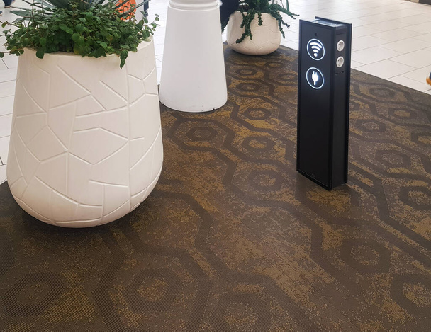 Zona Wi-Fi com um rack para carregar gadgets via USB e no aeroporto ou supermercado, plantas próximas em vasos de flores brancas. - Foto, Imagem