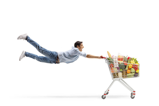 Full length shot of a casual young man flying with a shopping cart with food isolated on white background - Zdjęcie, obraz