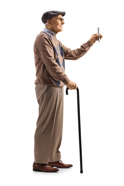 Full length profile shot of a grandfather standing with a cane and showing a smartphone isolated on white background - Zdjęcie, obraz
