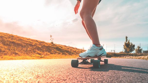 Pernas femininas em tênis brancos em um longboard dirigindo em uma estrada de asfalto em um dia ensolarado. - Foto, Imagem