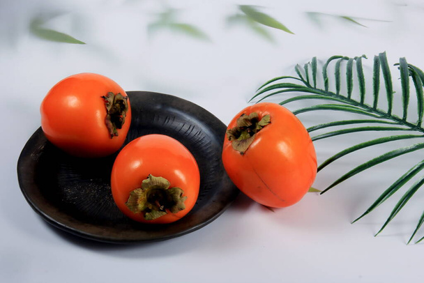 Three orange persimmon fruit on a black clay plate. Persimmon fruits in plate, light background with leaves, fresh hurma fruit  - Photo, Image
