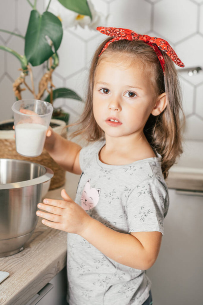 Koyu renk saçlı küçük bir kız. 3 yaşında, kırmızı bantlı. Mutfakta elmalı turta yapıyor. Çocuk ölçü kabından karıştırma kabına şeker döküyor. Çocuklar ev işlerine yardım eder. Çocuk yemek pişiriyor. Dikey - Fotoğraf, Görsel