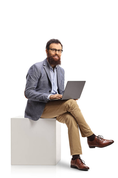 Bearded man sitting on a white cube and using a laptop computer isolated on white background - Foto, afbeelding