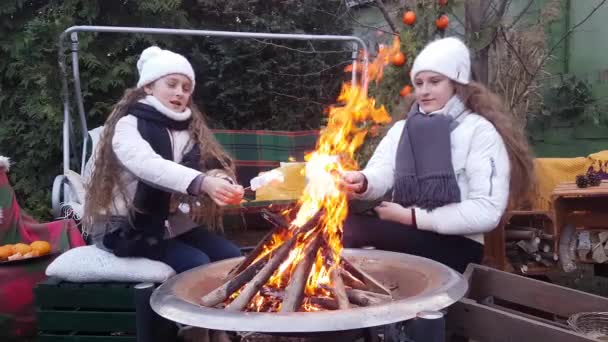 Kindermädchen mit langen weißen Locken in weißen Hüten und Jacken braten Marshmallows am Lagerfeuer im Garten - Filmmaterial, Video