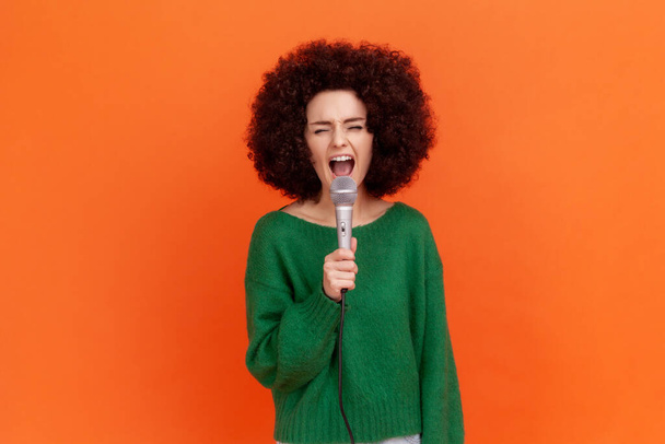 Portrait of attractive woman with Afro hairstyle wearing green casual style sweater singing loud favorite songs with microphone in hands, Indoor studio shot isolated on orange background. - Фото, зображення