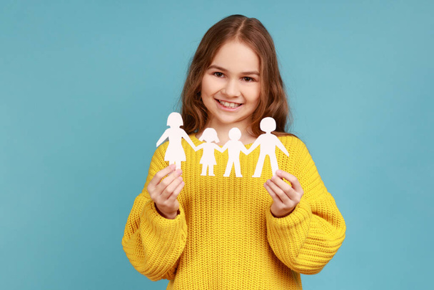 Cute little girl paper chain people which holding in hands, happy family, relationships, childhood, wearing yellow casual style sweater. Indoor studio shot isolated on blue background. - Fotografie, Obrázek