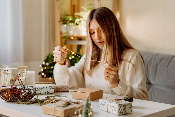 Donna che avvolge Natale eco scatole regalo naturali a casa. Preparazione regali su tavolo bianco con elementi decorativi, luci e oggetti Natale o Capodanno imballaggio fai da te Concetto - Foto, immagini