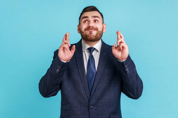 Portrait of hopeful man with beard wearing official style suit crossing fingers for luck, making a wish, dreaming of innermost, ritual. Indoor studio shot isolated on blue background. - Foto, Imagen