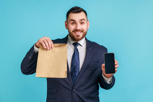 Happy man wearing official style suit holding empty display of cell phone and eco paper bag, online shopping, fast delivery, marketplace. Indoor studio shot isolated on blue background. - Zdjęcie, obraz