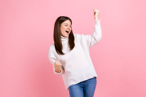Portrait of happy excited female standing with clenched fists, celebrating victory and her success, wearing white casual style sweater. Indoor studio shot isolated on pink background. - Foto, imagen