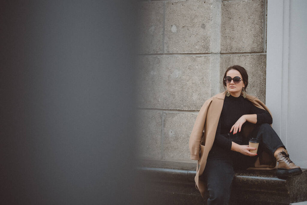 Portrait of a stylish woman dressed in a beige raincoat and sun goggles drinking a cup of coffee cloudy on the building. Street fashion concept. - Foto, afbeelding