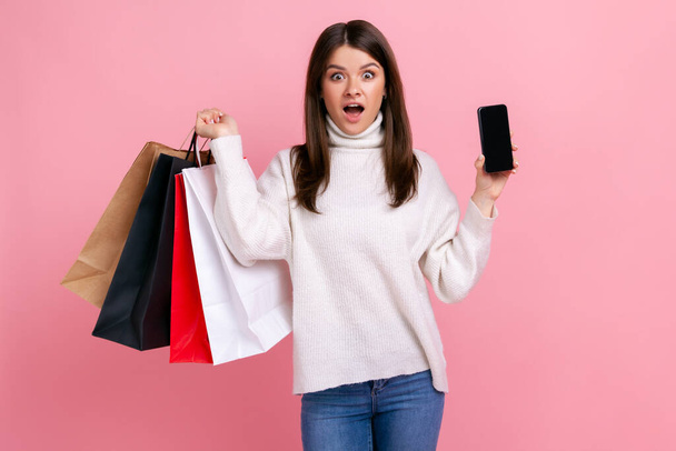 Mujer de pelo oscuro asombrada posando con bolsas de compras, mostrando el teléfono con pantalla en blanco para el anuncio, usando suéter de estilo casual blanco. Estudio interior plano aislado sobre fondo rosa. - Foto, Imagen