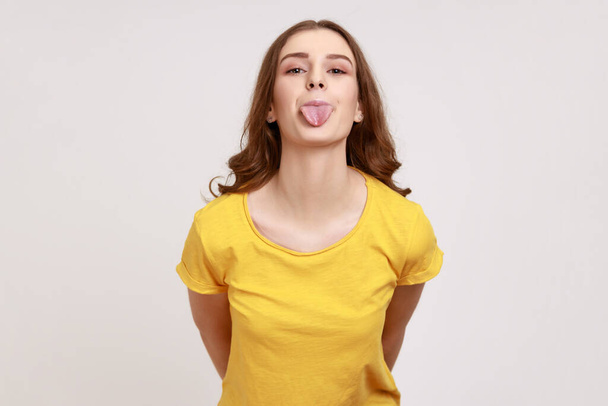 Young female with pleasant appearance wearing yellow T-shirt looking at camera with playful childish facial expression and showing tongue out. Indoor studio shot isolated on gray background. - Fotó, kép