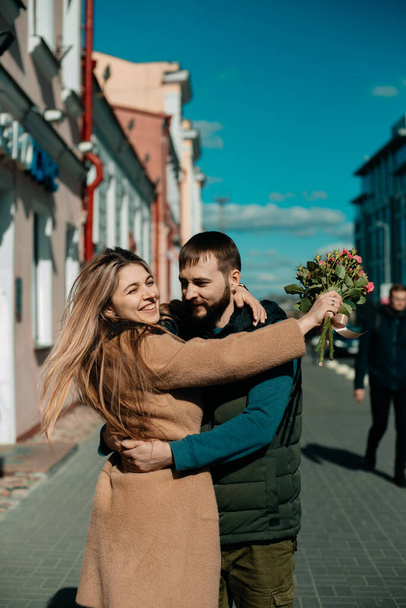 Portrait of two young people walking around the city in sunny weather. A happy girl with a bouquet of roses in her hands walks with a guy down the street. Date, love and care concept. - Zdjęcie, obraz