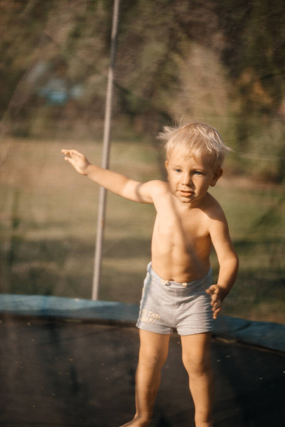 Cute little blond boy with blue eyes jumping on a trampoline outdoors on a sunny warm day. Outdoor activities, summer vacations. Happiness and fun concept. Selective focus. - Zdjęcie, obraz