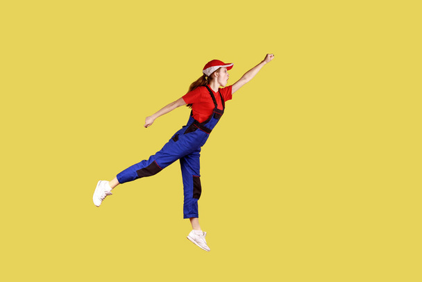 Side view portrait of woman worker flying to do her work like superhero, fast and high quality service, wearing work uniform and red cap. Indoor studio shot isolated on yellow background. - Photo, image