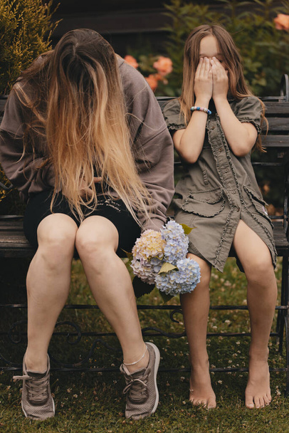 Two beautiful young girls with long hair posing on the bench with flowers in their hands. Happy girl enjoying the warm summer weather. Two sisters are having fun in nature. - Foto, imagen