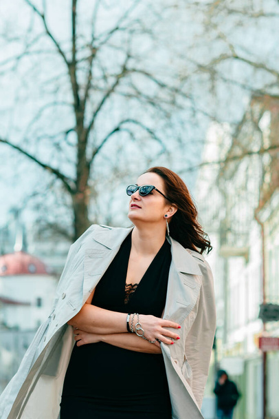 A stylish girl in a black dress in sunglasses, putting on a trench coat, walks through the city streets. Style and fashion concept. Portrait of a girl in sunglasses. - Photo, image